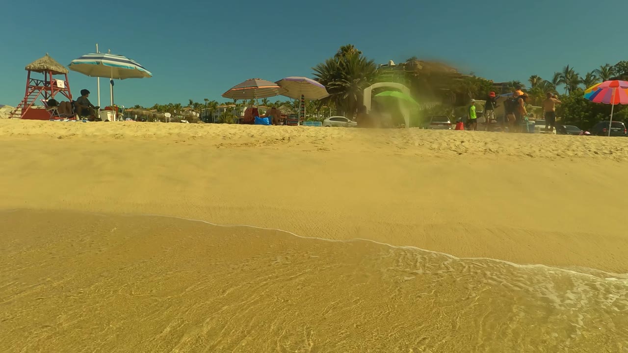 Sandy shoreline with beach umbrellas, people, and a small wave at the waterline.