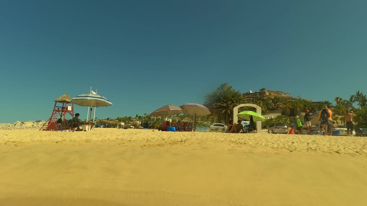 At 0:02 in San Jose del Cabo, a wider low-angle beach view places colorful umbrellas and scattered people across the middle of the frame, with parked cars, palms, and hillside behind them.