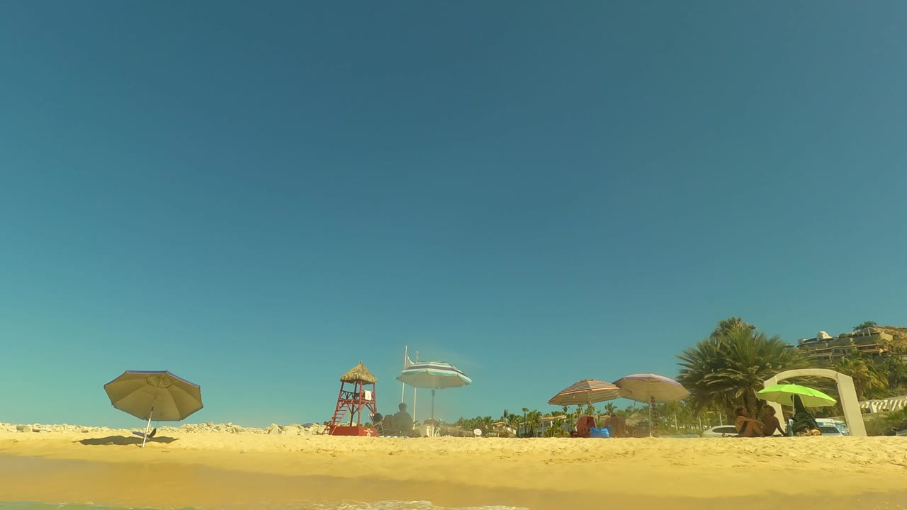 A low shoreline frame in San Jose Del Cabo shows wet sand, beach umbrellas, a red lifeguard tower, palms, and distant beachgoers under a clear blue sky.