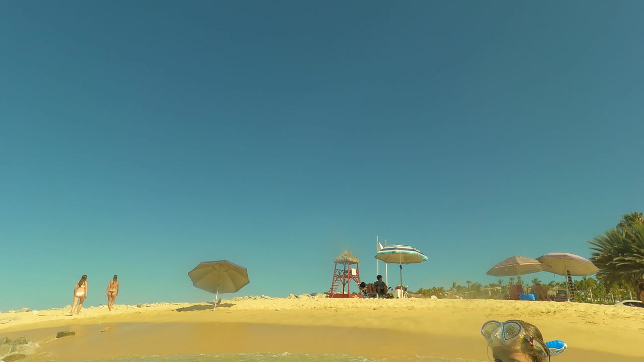 Two people walk on sandy beach near umbrellas and a lifeguard tower.