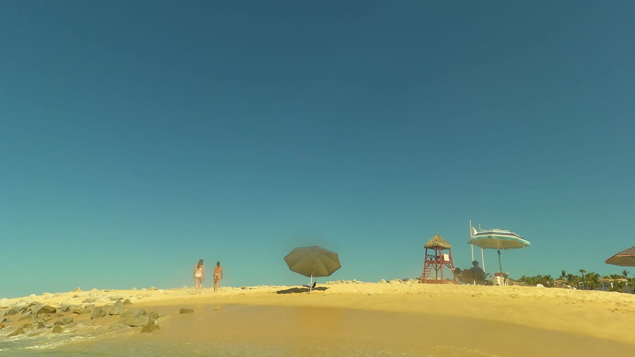 A low waterline view in San Jose Del Cabo shows a person partly visible at the bottom edge, with sandy beach, umbrellas, palms, and a lifeguard tower in the background.