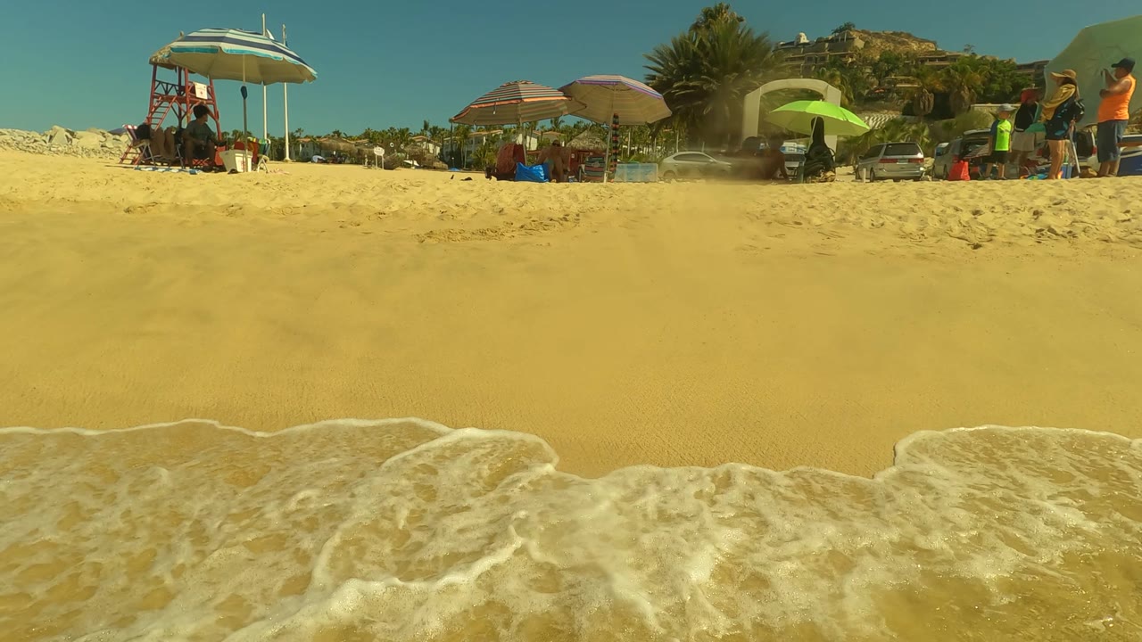 Low shoreline frame in San Jose del Cabo with foamy water crossing yellow sand, beach umbrellas, seated people, and a hillside beyond the public beach.