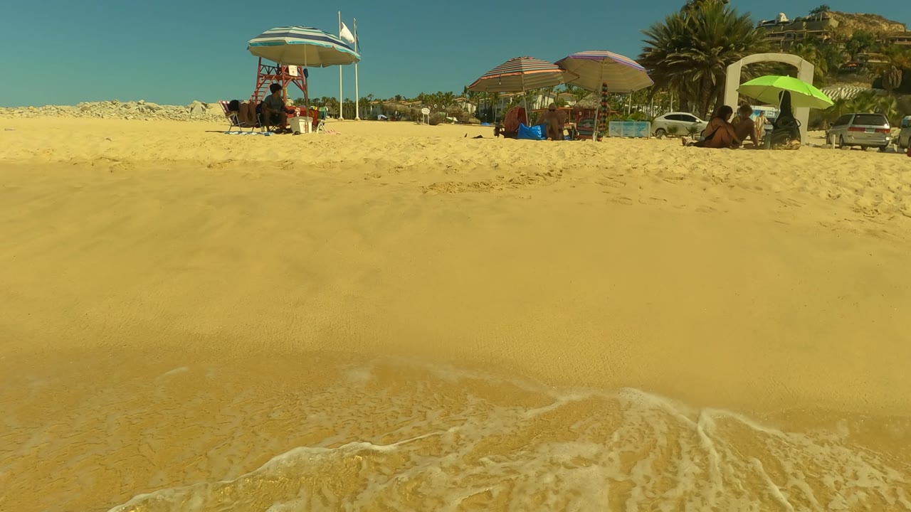 Wide sandy beach with small waves, umbrellas, people, vehicles, and hillside houses.
