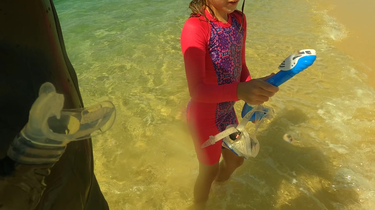 A child in red swimwear stands in clear shallow water in San Jose del Cabo, holding blue snorkeling gear while waves wash around their legs.