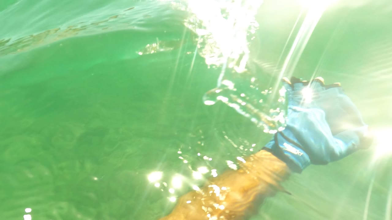 A blue-gloved hand and tan forearm move near the bright water surface in San Jose del Cabo, with bubbles, sun flare, and green water below, no visible people.
