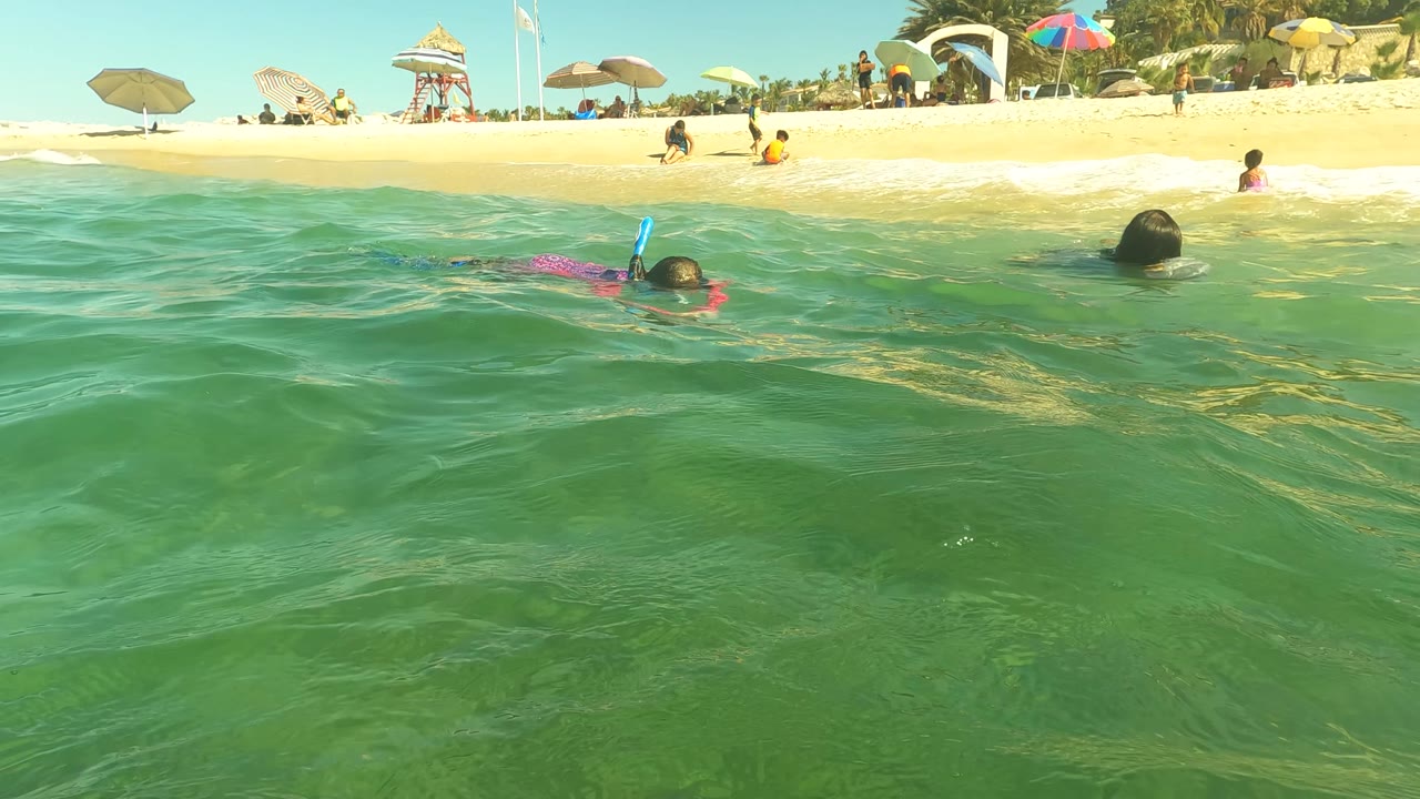 Water-level view in San Jose del Cabo with a snorkeler in shallow green ocean, beach umbrellas on sand, and several people visible near the shoreline.