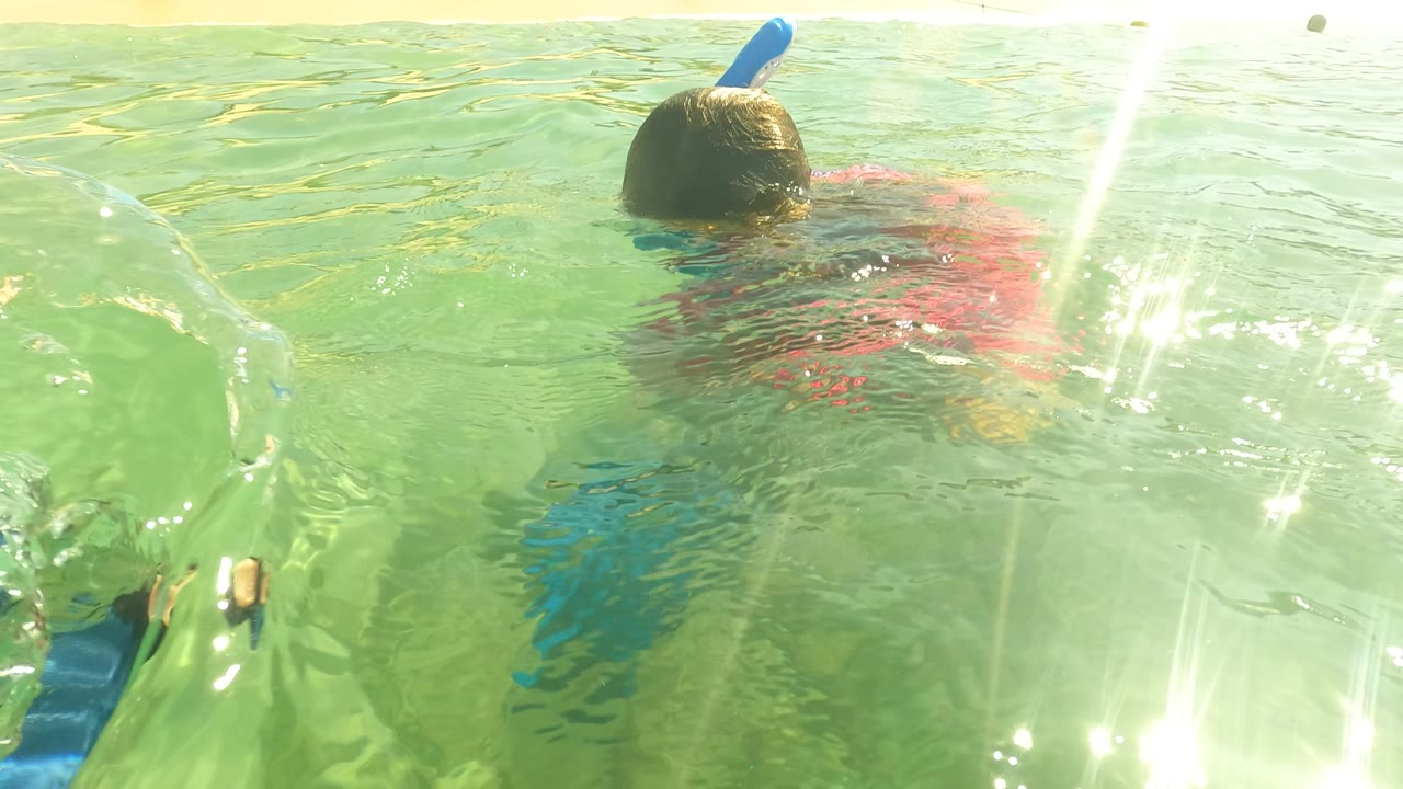 At San Jose del Cabo, a child with a blue snorkel moves away from the camera in shallow green ocean water, with sun glare on the surface and beachgoers faintly visible along the shore.