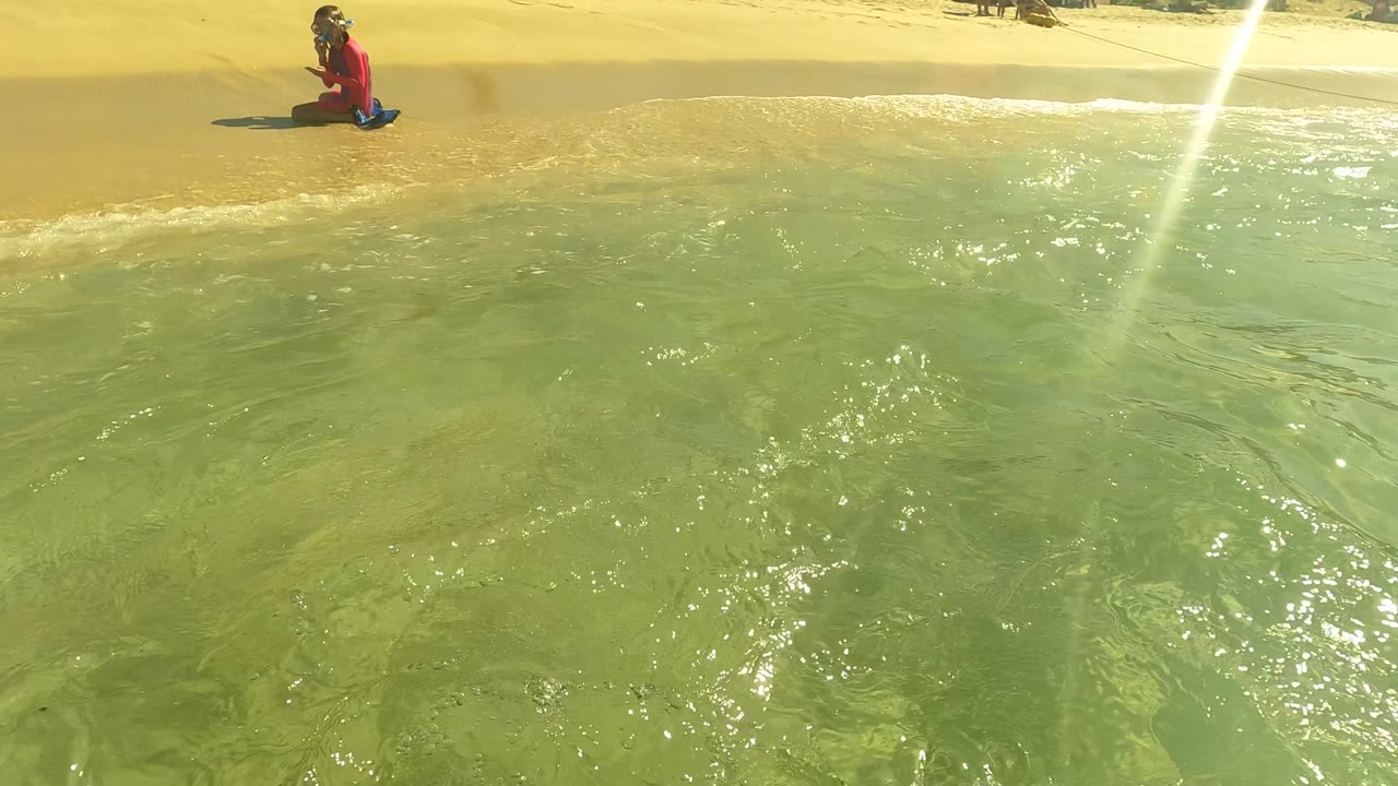 At San Jose del Cabo, a child kneels on the wet sand near shallow green water, with bright glare on the ocean surface and beach visible behind.