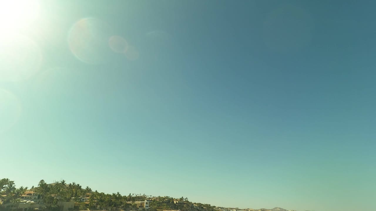 The camera view lifts toward bright sun flare and blue sky over the San Jose del Cabo shoreline, with a thin strip of beach, trees, and ocean at the bottom.
