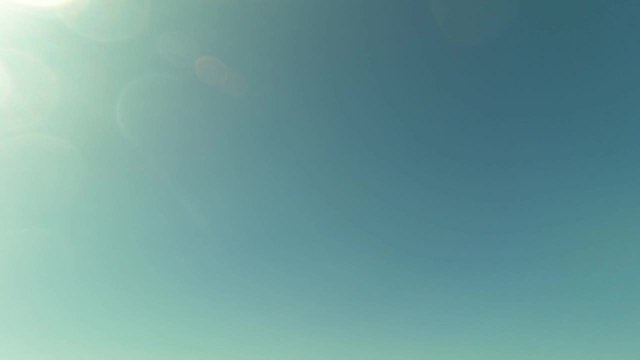 A wide blue sky dominates the frame above San Jose del Cabo, with soft sun flare near the upper left and only a low band of ocean and shoreline visible.