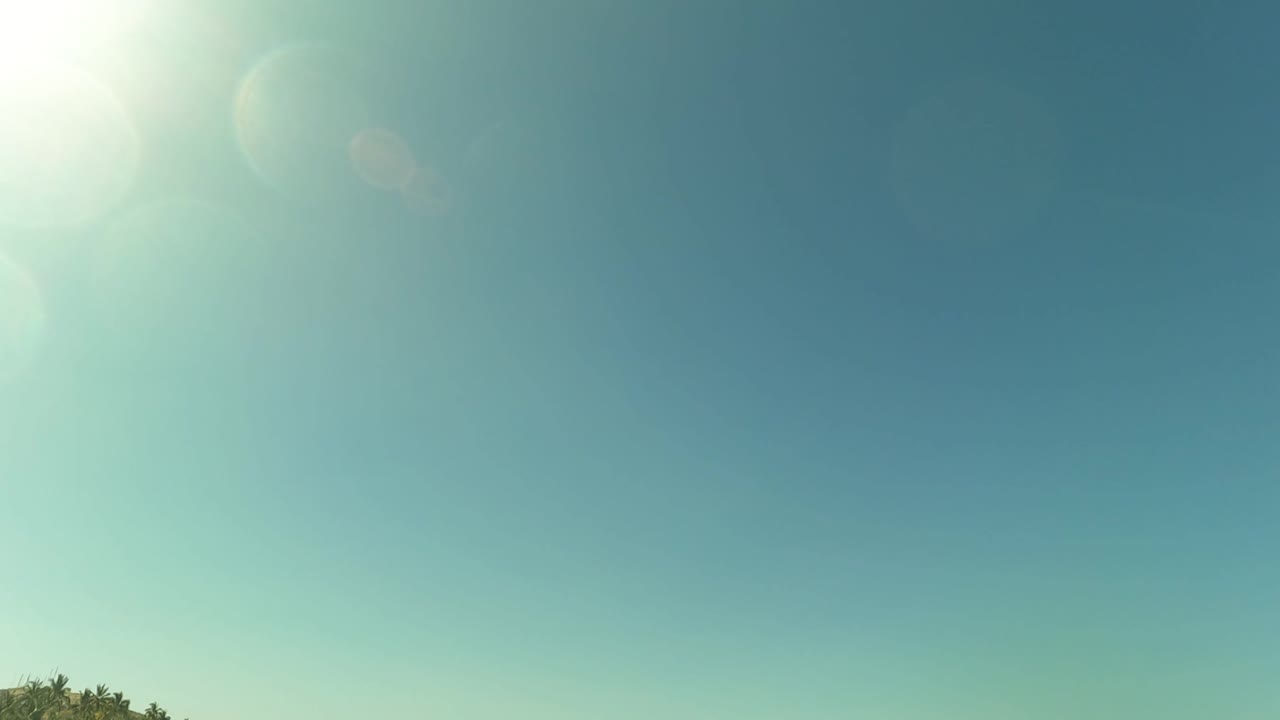The frame holds mostly bright blue sky and pale sun flare, with the San Jose del Cabo shoreline barely visible along the bottom edge and no visible people.