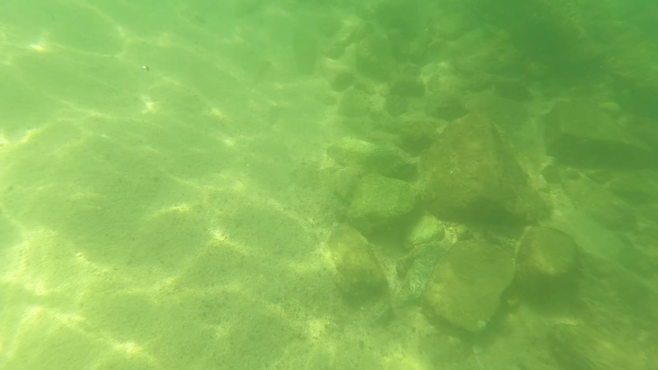 Opening underwater view in San Jose del Cabo, Mexico, showing pale sand, scattered rounded rocks, green water, bright caustic light, and no visible people.