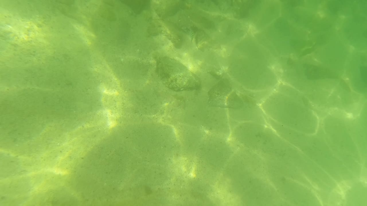 Mid-clip underwater view in San Jose del Cabo, Mexico, showing scattered stones through green-tinted water and bright shifting reflections over the sandy bottom, with no visible people.