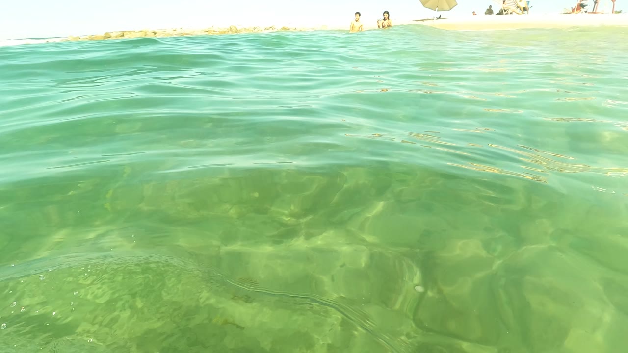 Low waterline view in San Jose del Cabo, Mexico, with rippled green water in the foreground and a sandy beach with distant people and umbrellas behind it.