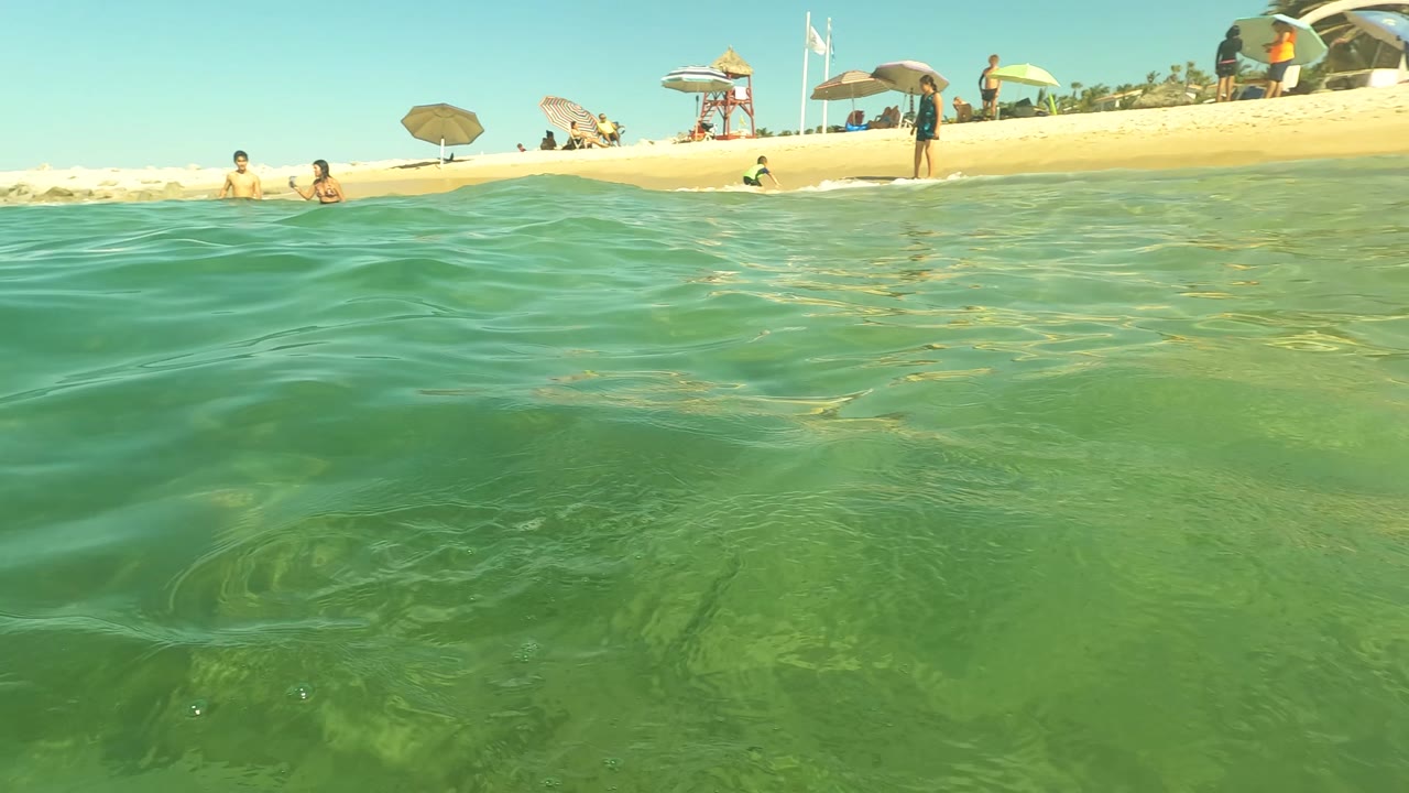 Water-level view in San Jose del Cabo, Mexico, looking across green surface water toward a sandy beach with umbrellas, beachgoers, and swimmers near shore.