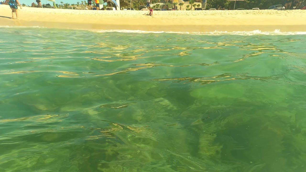 Water-level frame in San Jose Del Cabo, Baja California Sur, with green-blue shallow water in front and small distant beachgoers along the sand in the background.