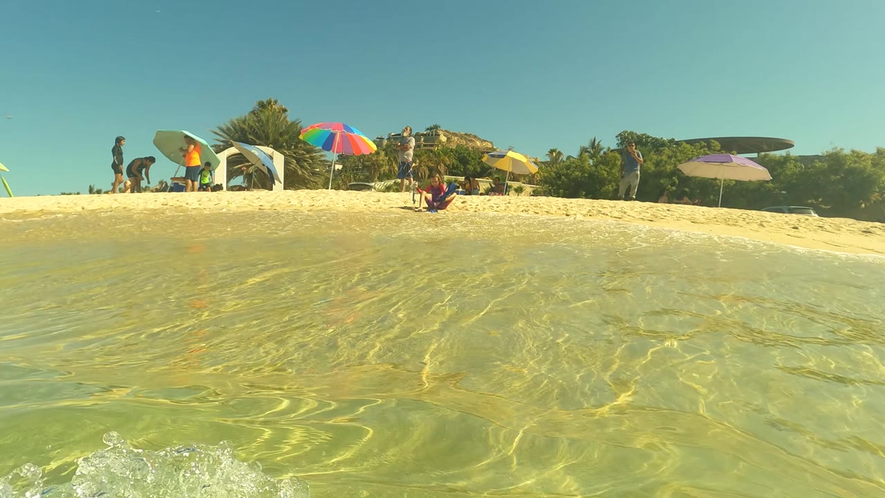 Clear green water ripples across the foreground at a San Jose Del Cabo beach, with beachgoers, colorful umbrellas, surfboards, shrubs, and bright sky beyond the sand.