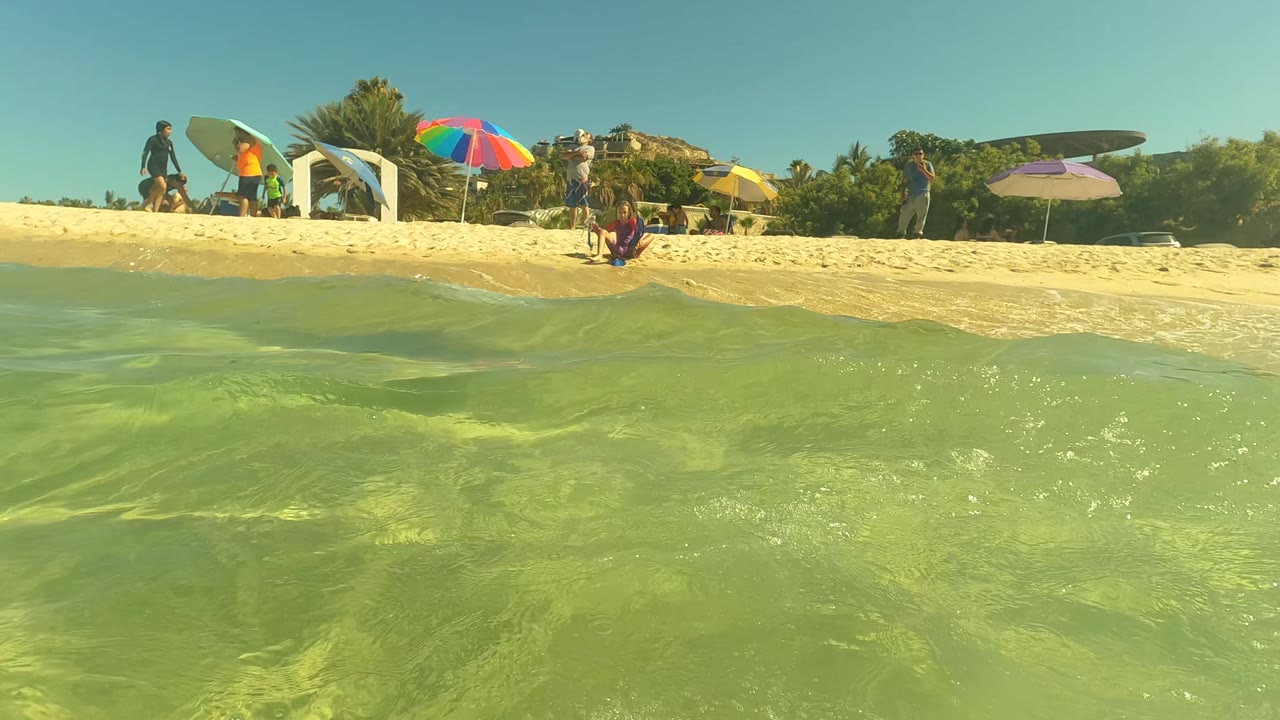 Shallow green water in the foreground with beachgoers, umbrellas, and surfboards on sand behind it.
