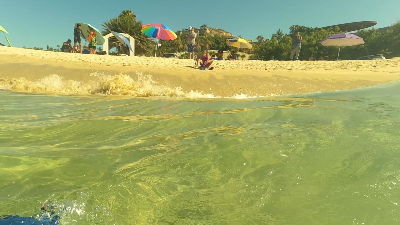 A small shore wave rises across clear green water in San Jose Del Cabo, with a sandy beach, seated and standing beachgoers, umbrellas, and surfboards visible along the horizon.