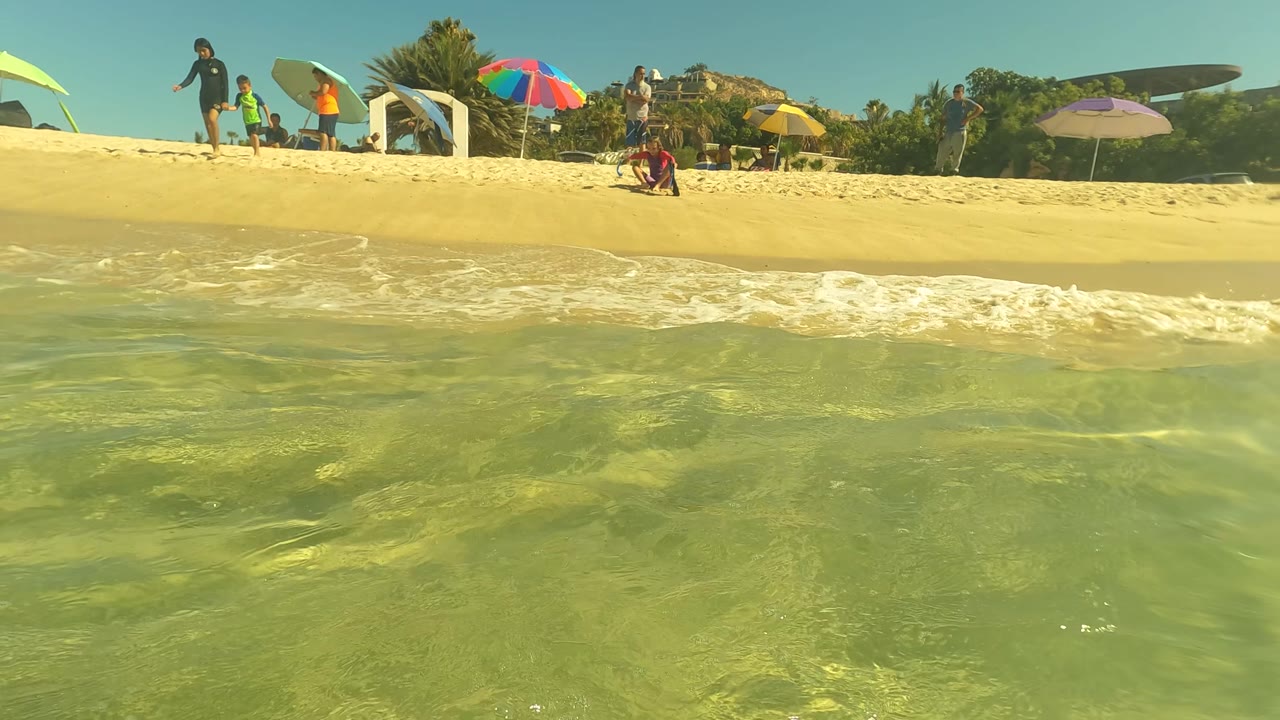 Low waterline frame in San Jose del Cabo showing clear shallow water in front of a sandy beach with umbrellas, boards, and people along the shore.