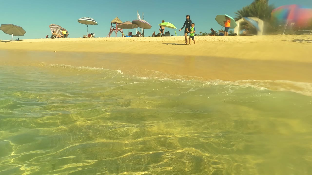 San Jose del Cabo shoreline frame with sunlit shallow water, a sandy beach band, colorful umbrellas, and small figures visible near the surf line.