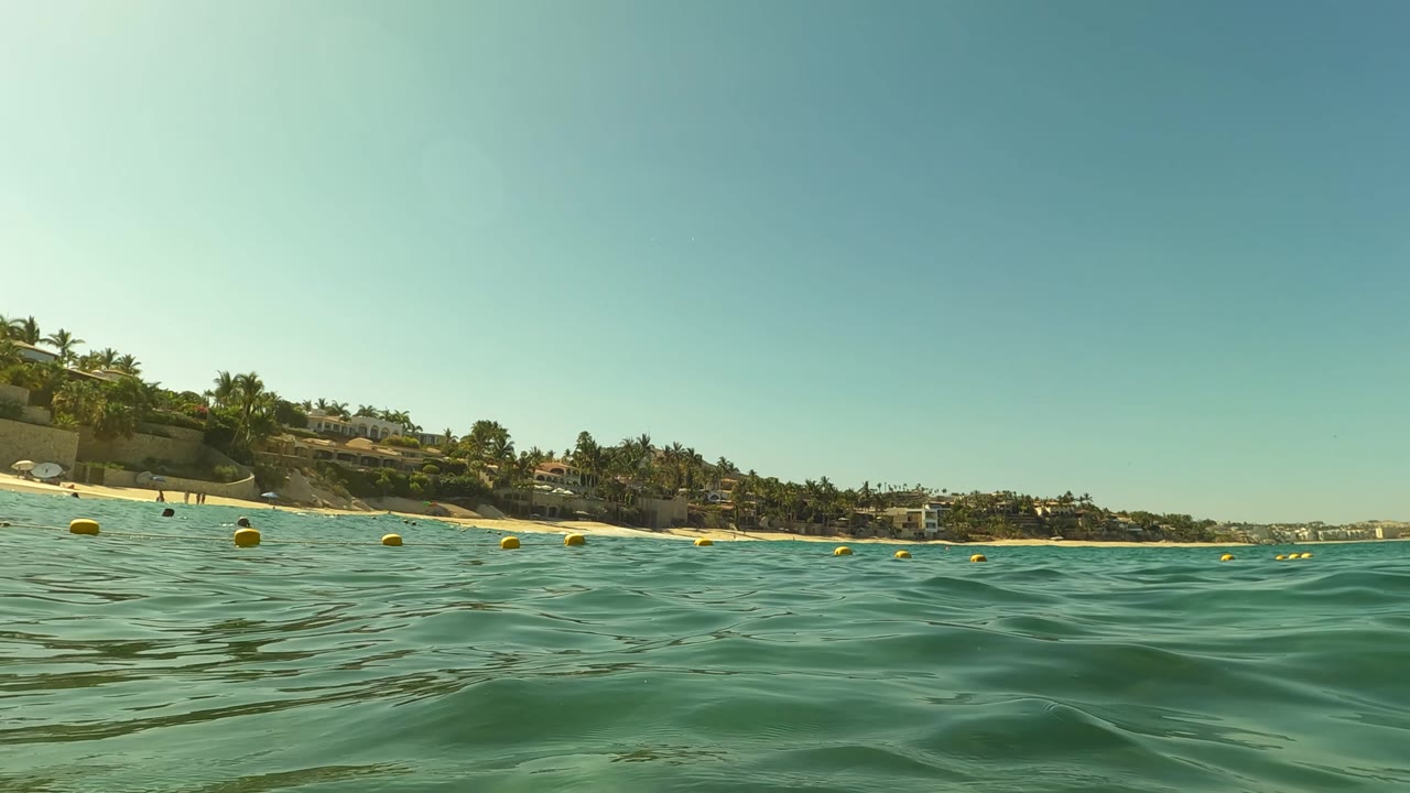 Ocean surface with yellow buoys and beach buildings in the distance.