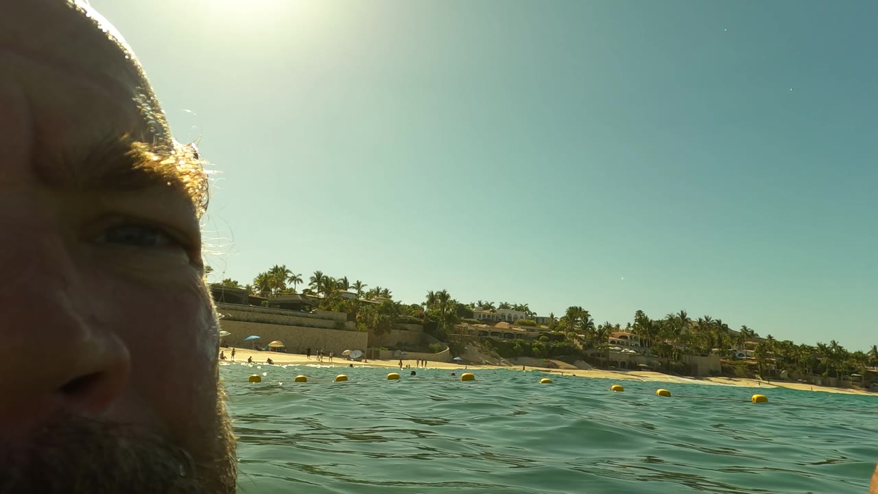 Close partial view of a bearded person’s face at the edge of the frame in San Jose del Cabo, with ocean water, yellow buoys, shoreline trees, and buildings behind.