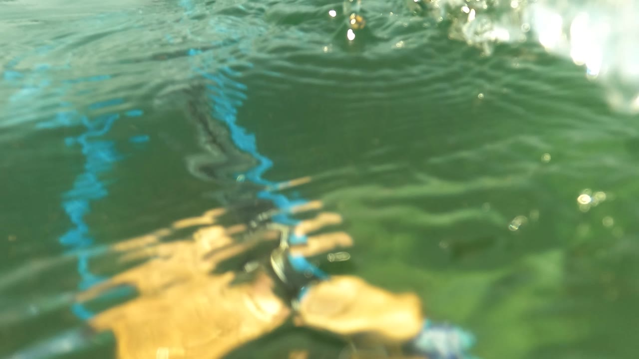 At 0:02 in San Jose del Cabo, a hand sits near a blue hose and dark metal equipment under bright rippled water, with green water filling the frame and no visible faces.
