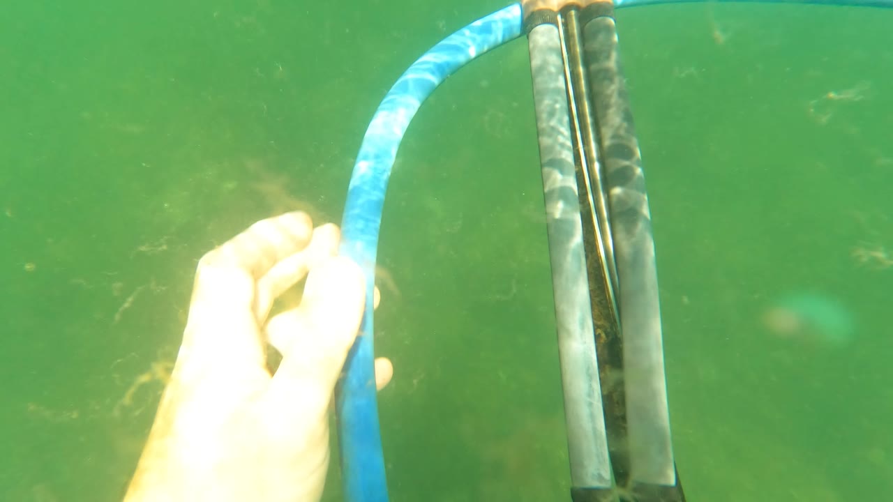 A hand reaches toward the blue hose next to vertical speargun rods in green water near San Jose Del Cabo, Mexico, with no face visible.