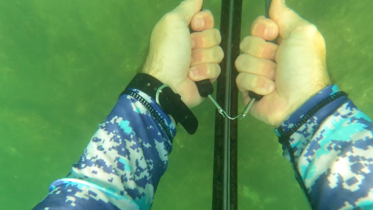 Two hands grip the drawn speargun bands beside the black shaft in San Jose Del Cabo green water, with bright underwater light and no visible faces.