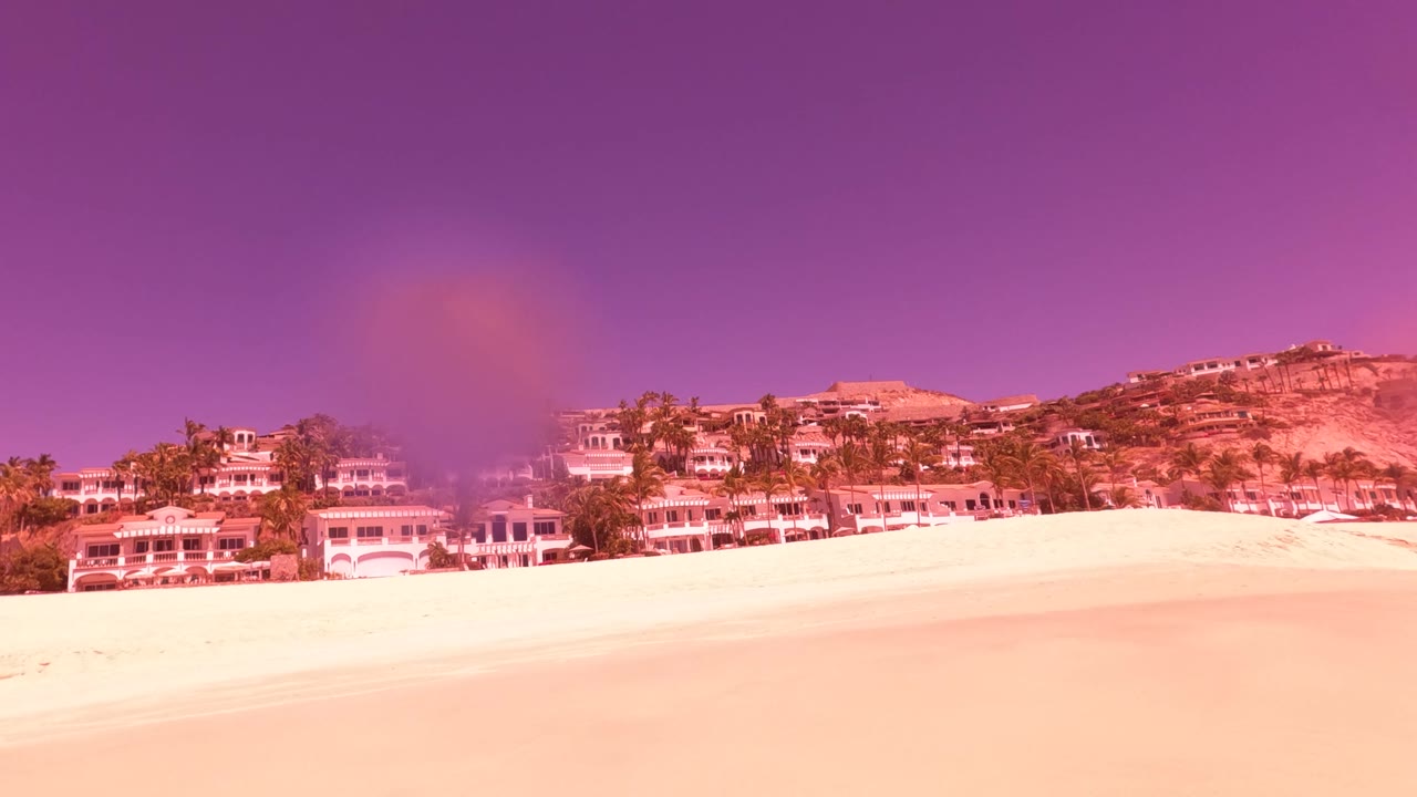 Wide red and magenta tinted view of a sandy San Jose Del Cabo beach with resort buildings and palms above the shore, surf at the far right, and no visible people.