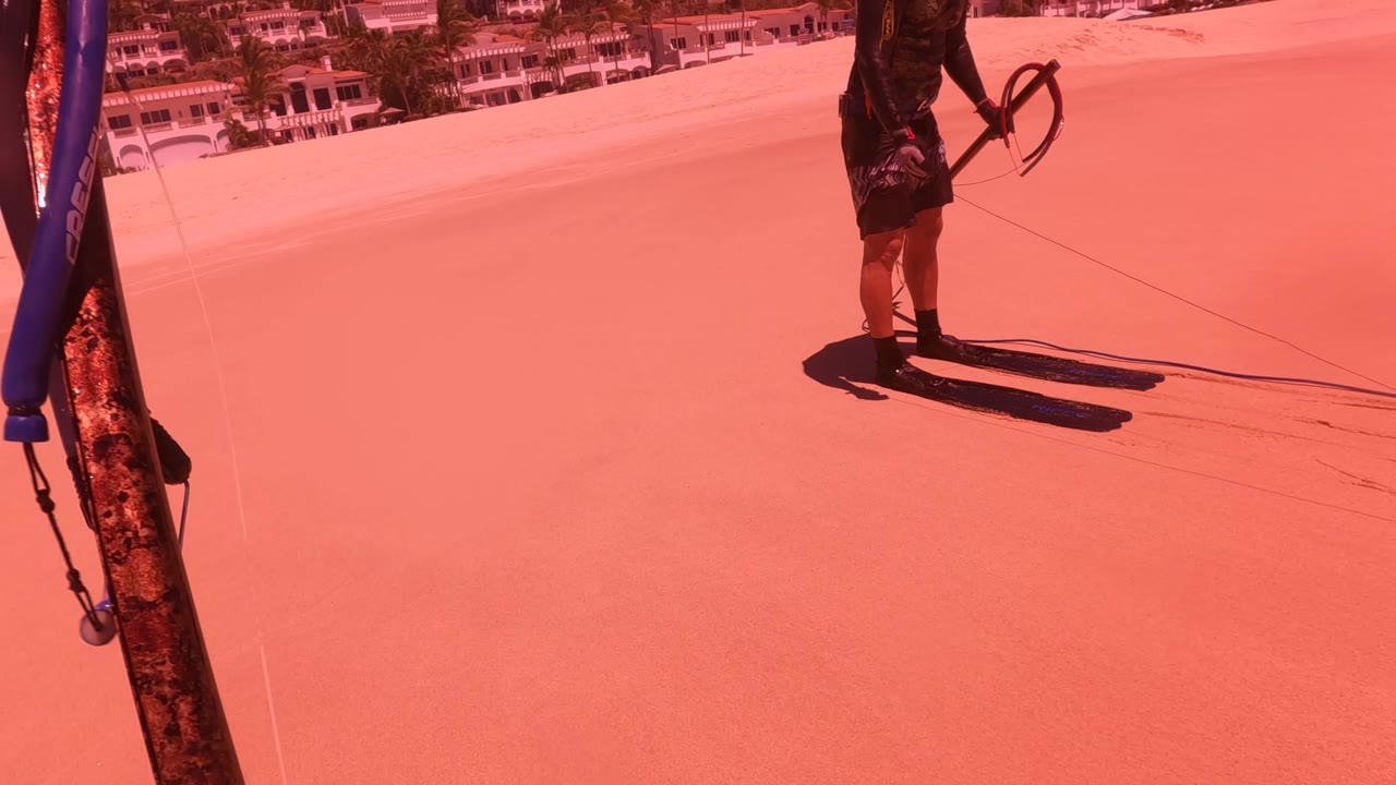 A person stands on a red-tinted sandy slope in San Jose del Cabo with a rope in hand, hillside buildings in the background, and part of a pole or rig at the left edge.