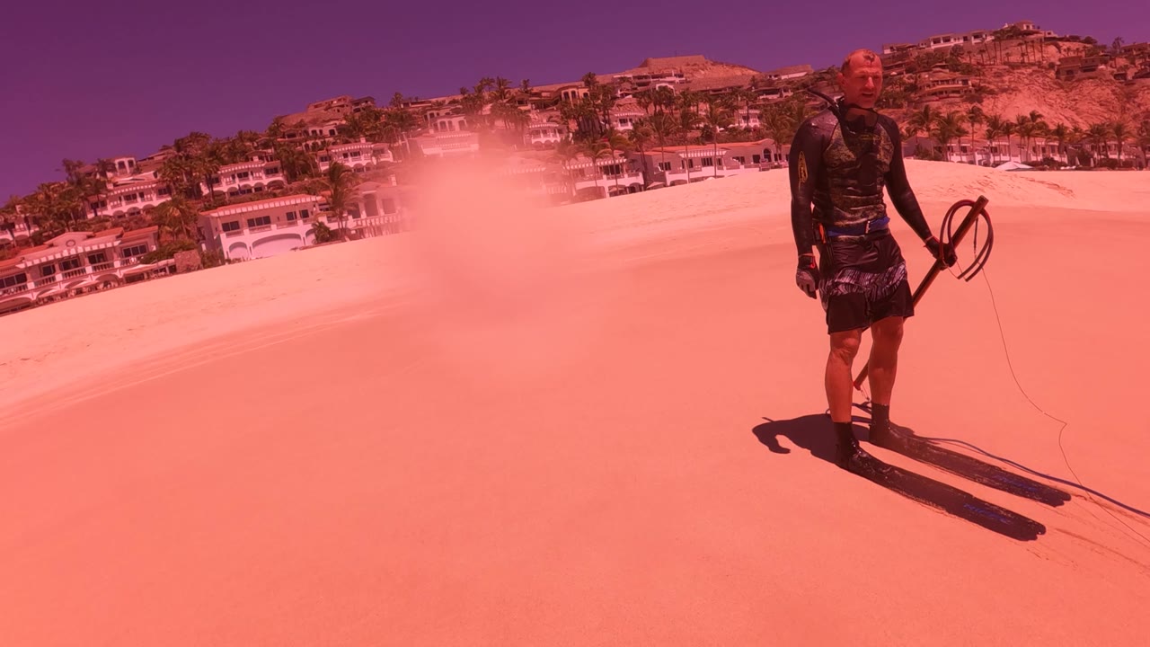 A person stands on the sandy incline in San Jose del Cabo while a pale dust cloud lifts across the slope and hillside buildings remain visible behind the sand.