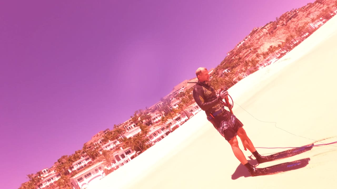 Kite surfer standing on a bright sandy beach in San Jose del Cabo, Mexico, with control lines in hand and beach buildings in the background.
