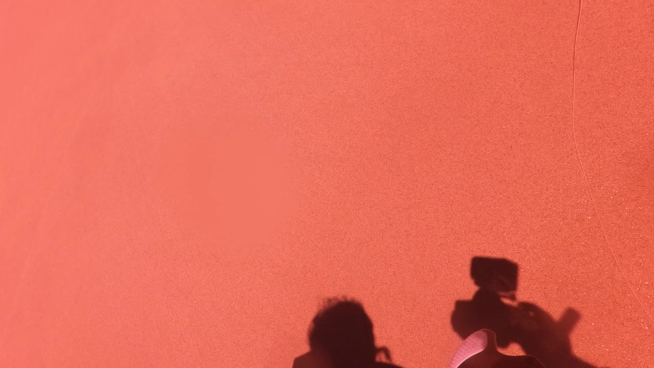 Pink-orange sand fills the frame in San Jose del Cabo, Mexico, with the shadow of a camera operator and a small portion of gear visible at the right edge.