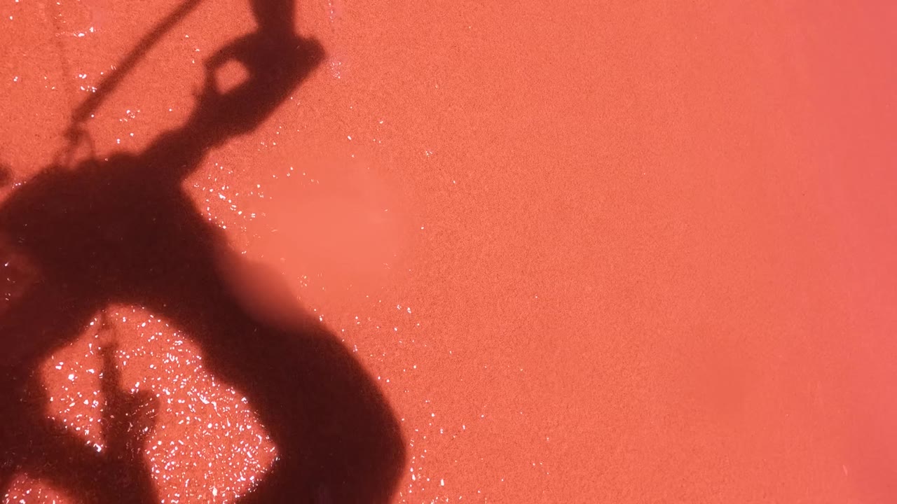 A dark shadow of a paddleboarder holding a paddle falls across pink salt water in San Jose Del Cabo, with bright specks on the surface and no face visible.