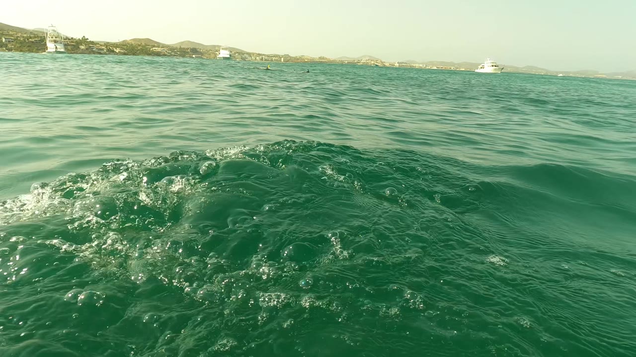 Surface-level San Jose del Cabo ocean frame with a patch of bubbling water in the foreground, distant boats, coastline, and no visible people.