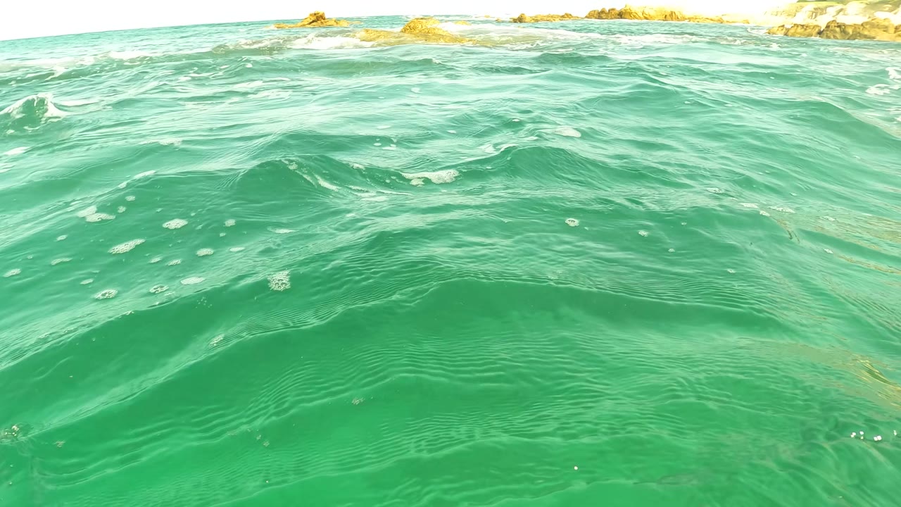 Water-level view of turquoise ocean in San Jose Del Cabo with small waves, foam flecks, distant rocky outcrops, and no visible people.