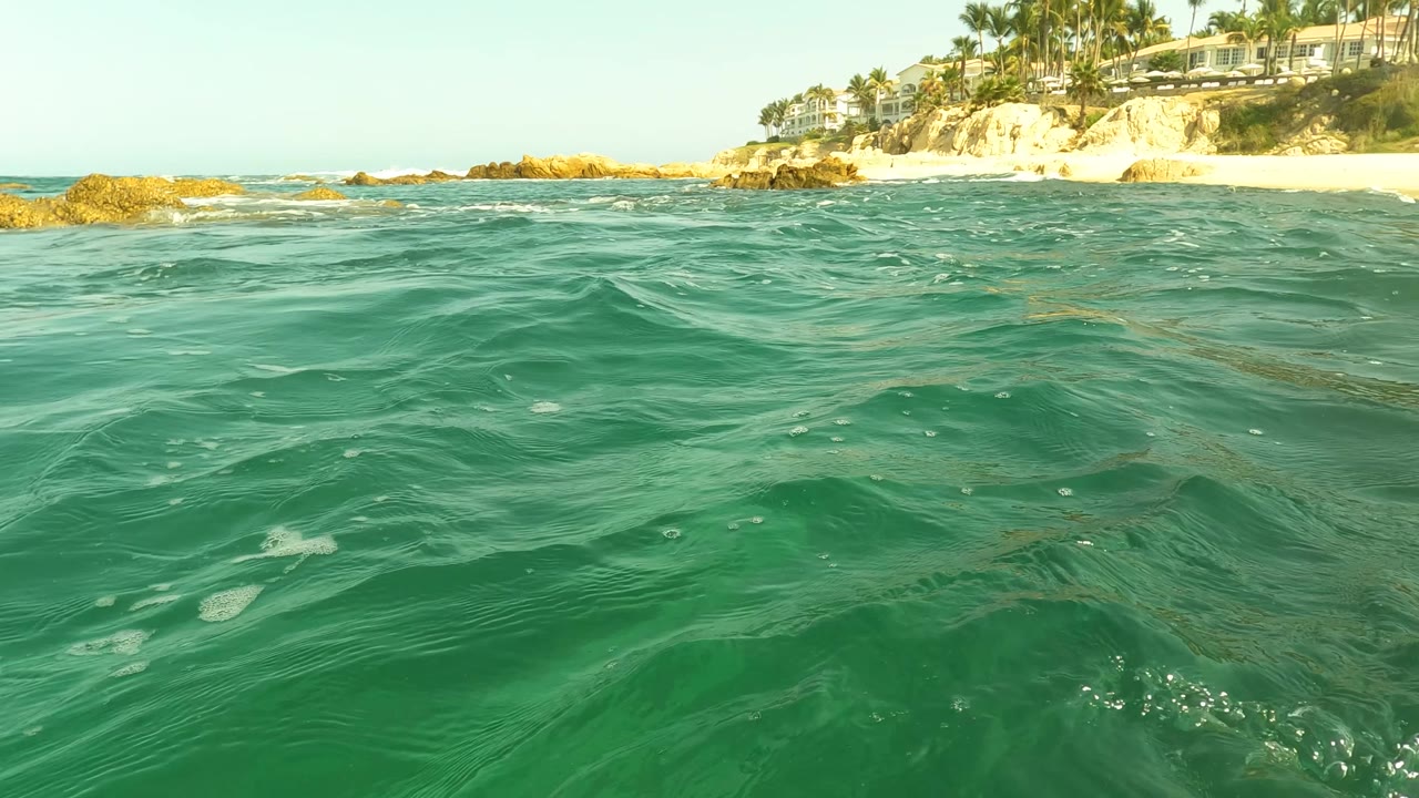 Water-level frame in San Jose Del Cabo showing rippled green water, rocky shoreline, beach, hillside buildings, palms, and no visible people.