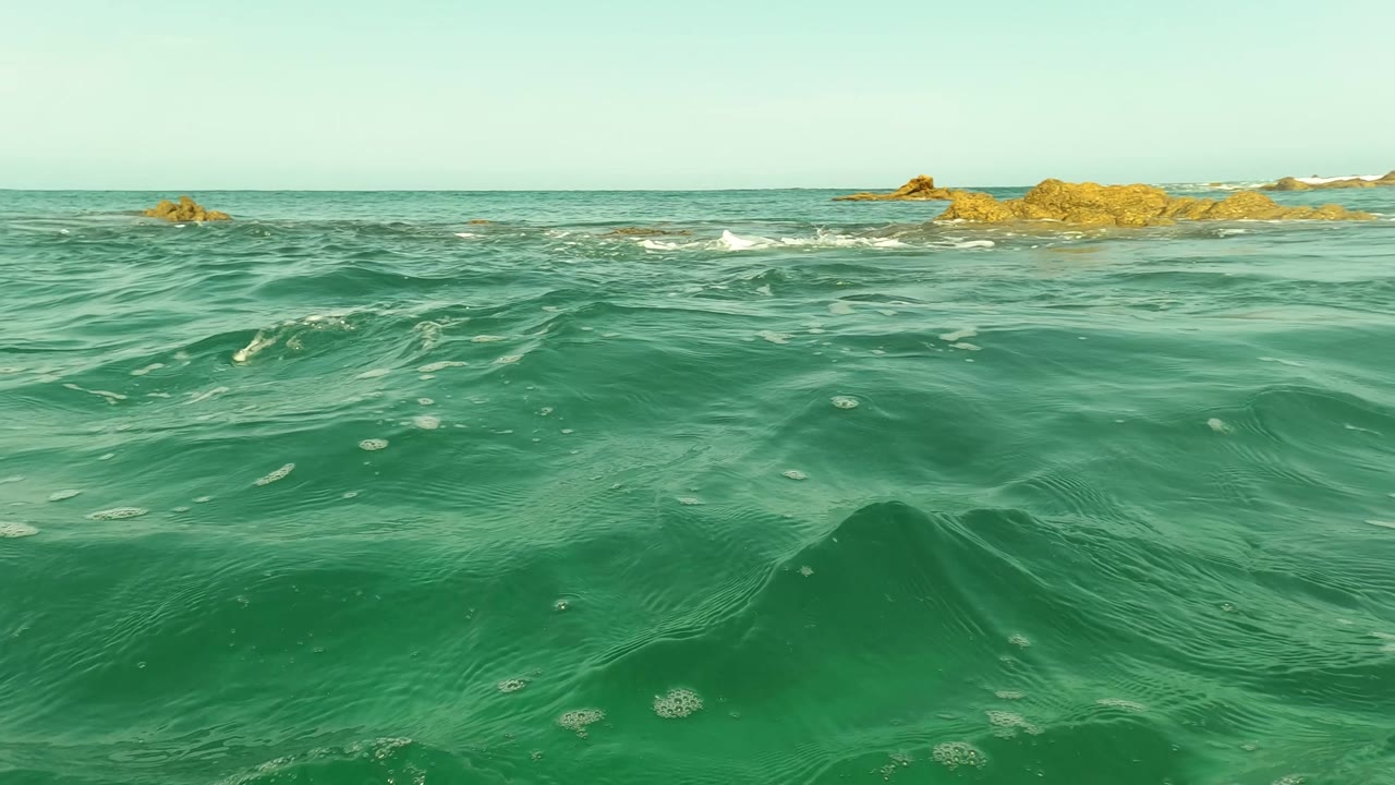 Low surface view of a darker turquoise wave in San Jose Del Cabo with rocky outcrops on the horizon and no visible people.