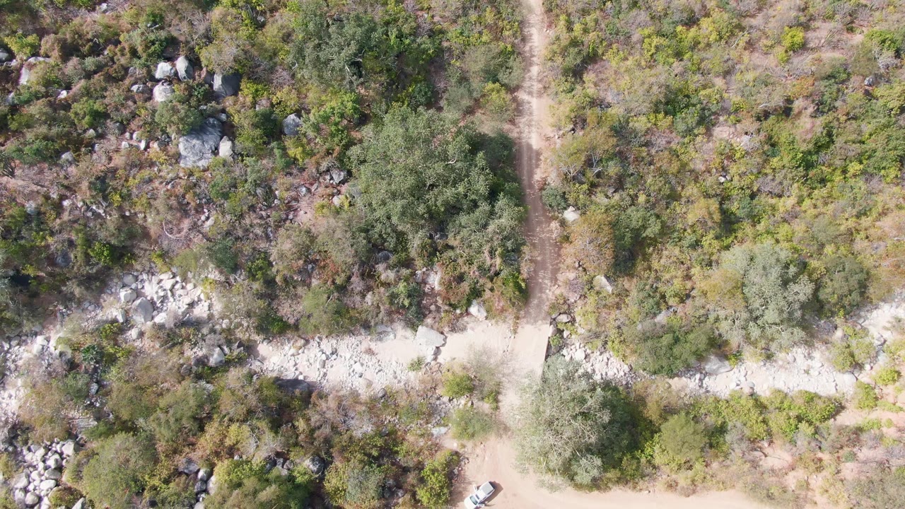 Top-down San Dionisio frame showing rocky scrubland, a light dirt road across the lower frame, a vertical trail, and no visible people.