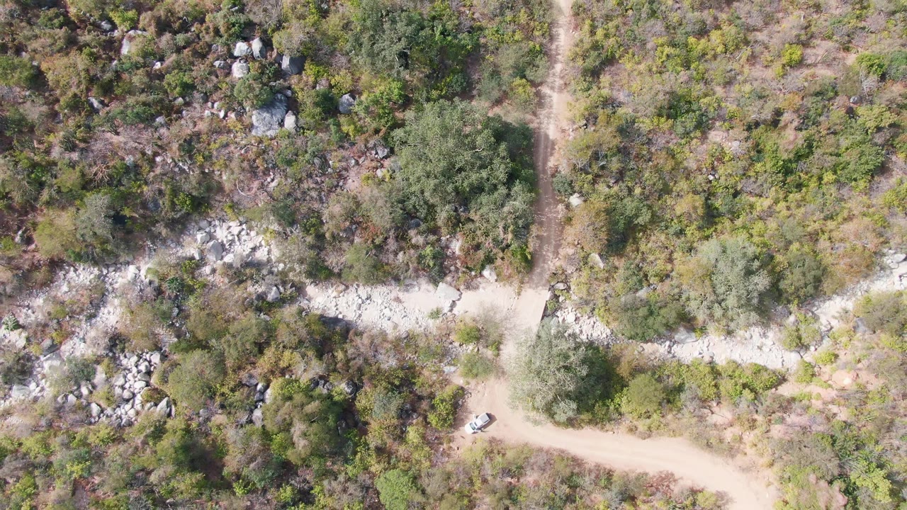 Aerial view in San Dionisio, Baja California Sur, showing the vehicle near a bend in the pale road, with dense scrub, scattered trees, and light-colored rock outcrops around it, and no visible people.