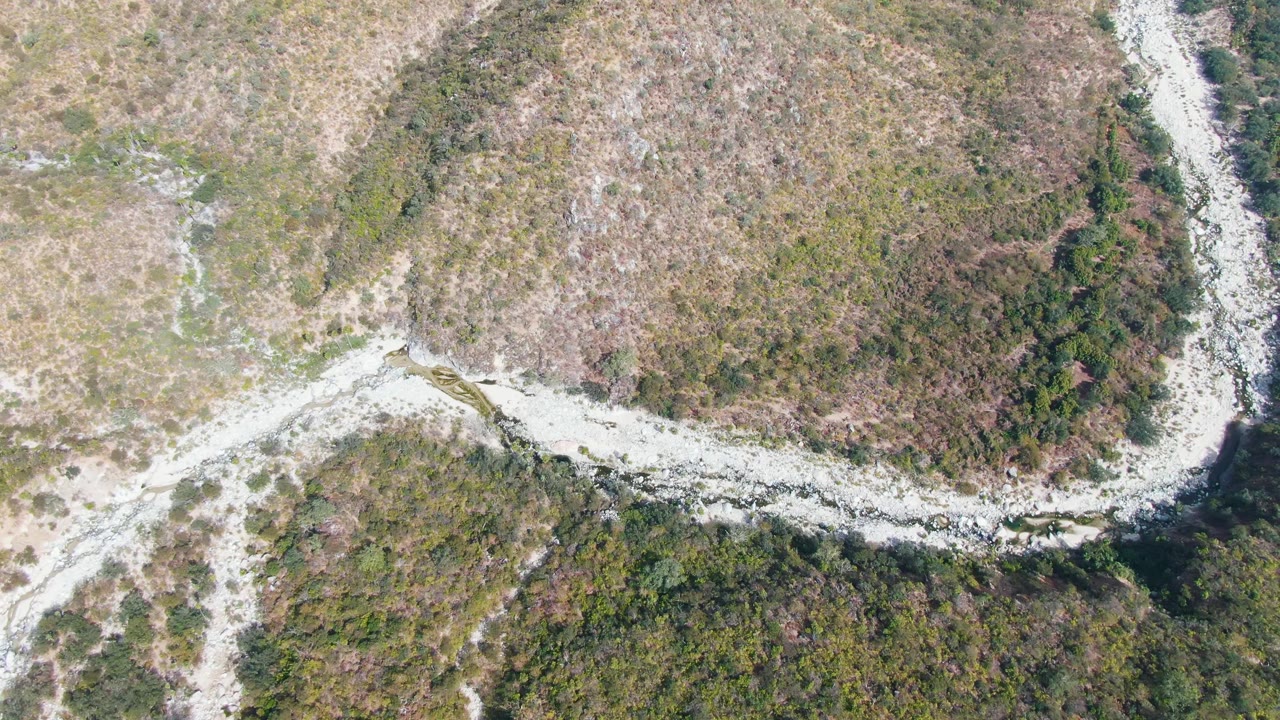 Aerial frame from San Dionisio, Baja California Sur showing the road crossing a light rocky channel between dry brush above and denser green forest below, with no visible people.