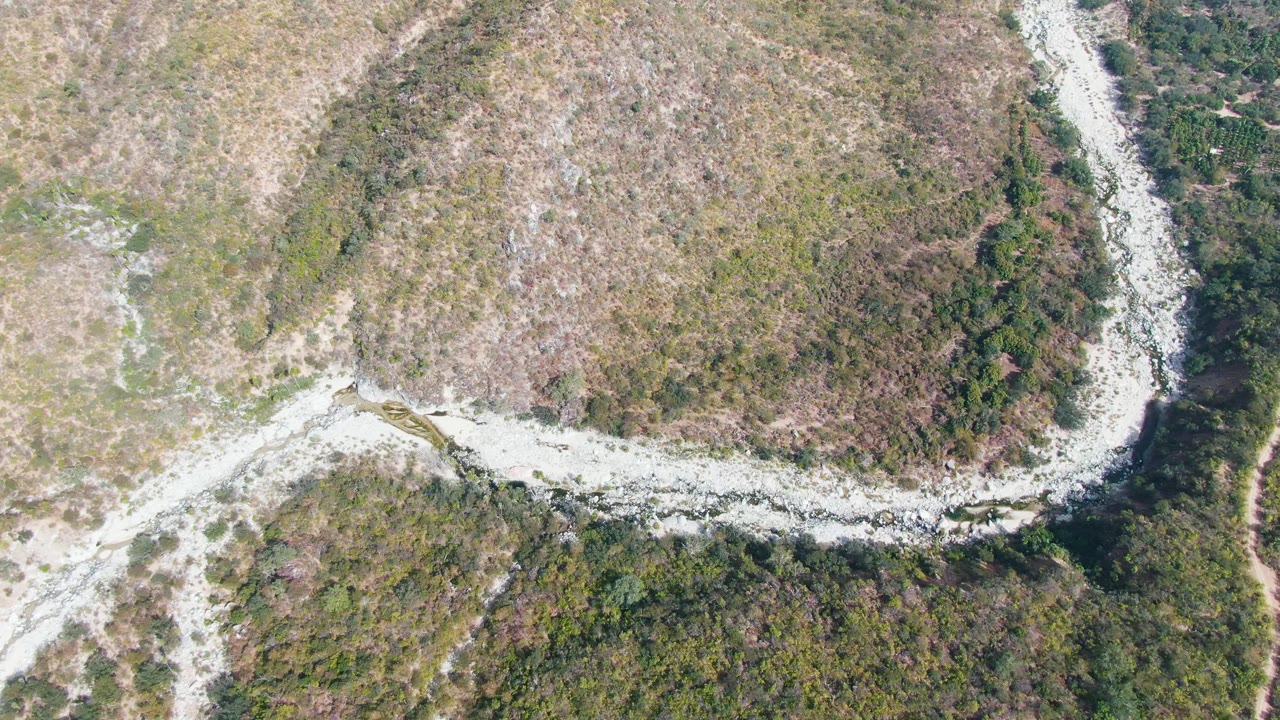 Aerial frame in San Dionisio, Baja California Sur with the rocky road curve more centered between dry scrub and compact green tree cover, with no visible people.