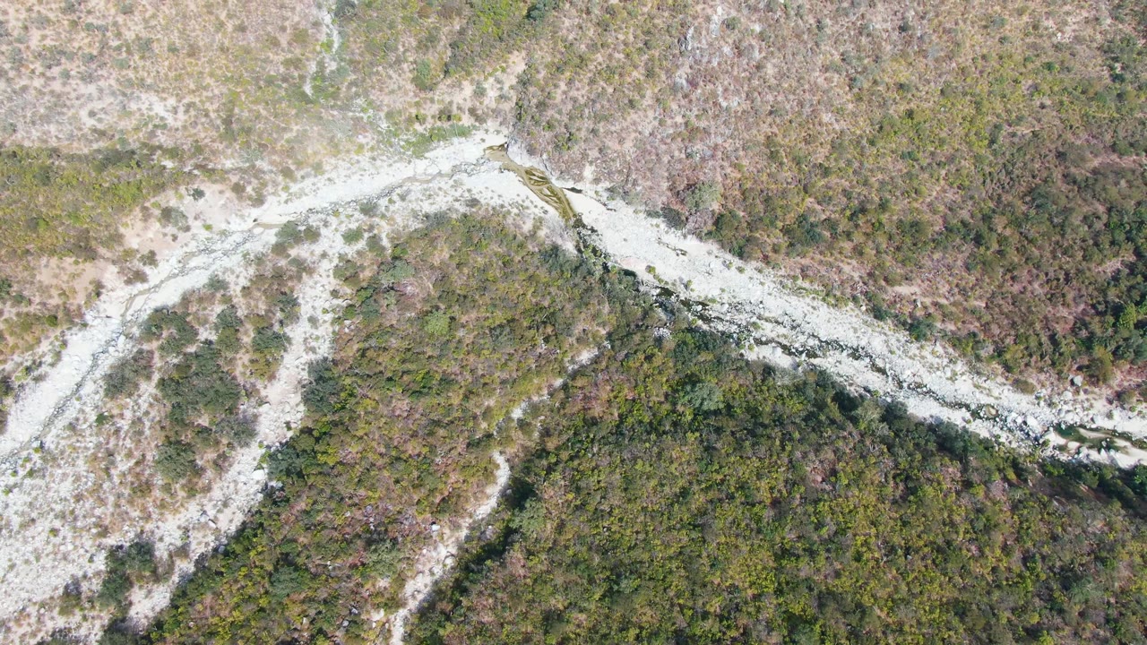 Aerial frame over San Dionisio terrain with the pale path running diagonally through denser green brush below and lighter open ground above, with no visible people.
