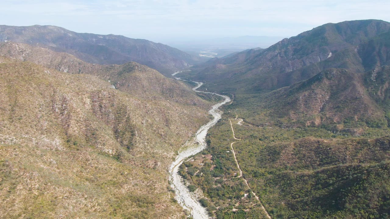 Aerial frame over San Dionisio, Baja California Sur showing a pale winding riverbed, dry brown slopes, green valley vegetation, distant mountains, and no visible people.