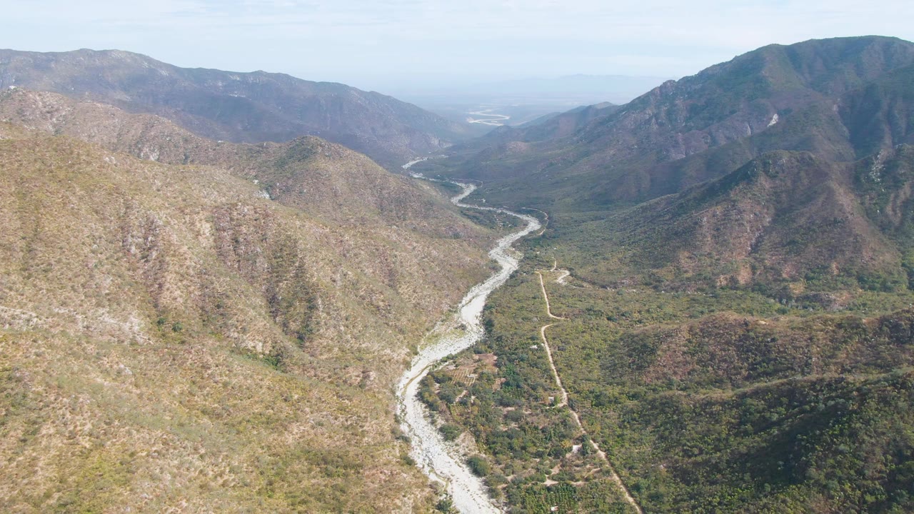 Aerial frame in San Dionisio, Baja California Sur with the riverbed centered lower in the valley, a pale rocky slope at left, darker mountains at right, and no visible people.