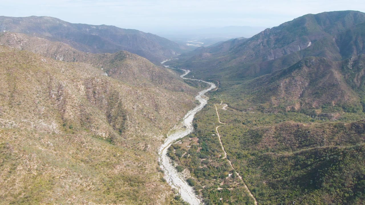 Aerial frame over San Dionisio, Baja California Sur showing a curving pale riverbed, scrub-covered valley floor, broad brown hillsides, layered mountains, and no visible people.
