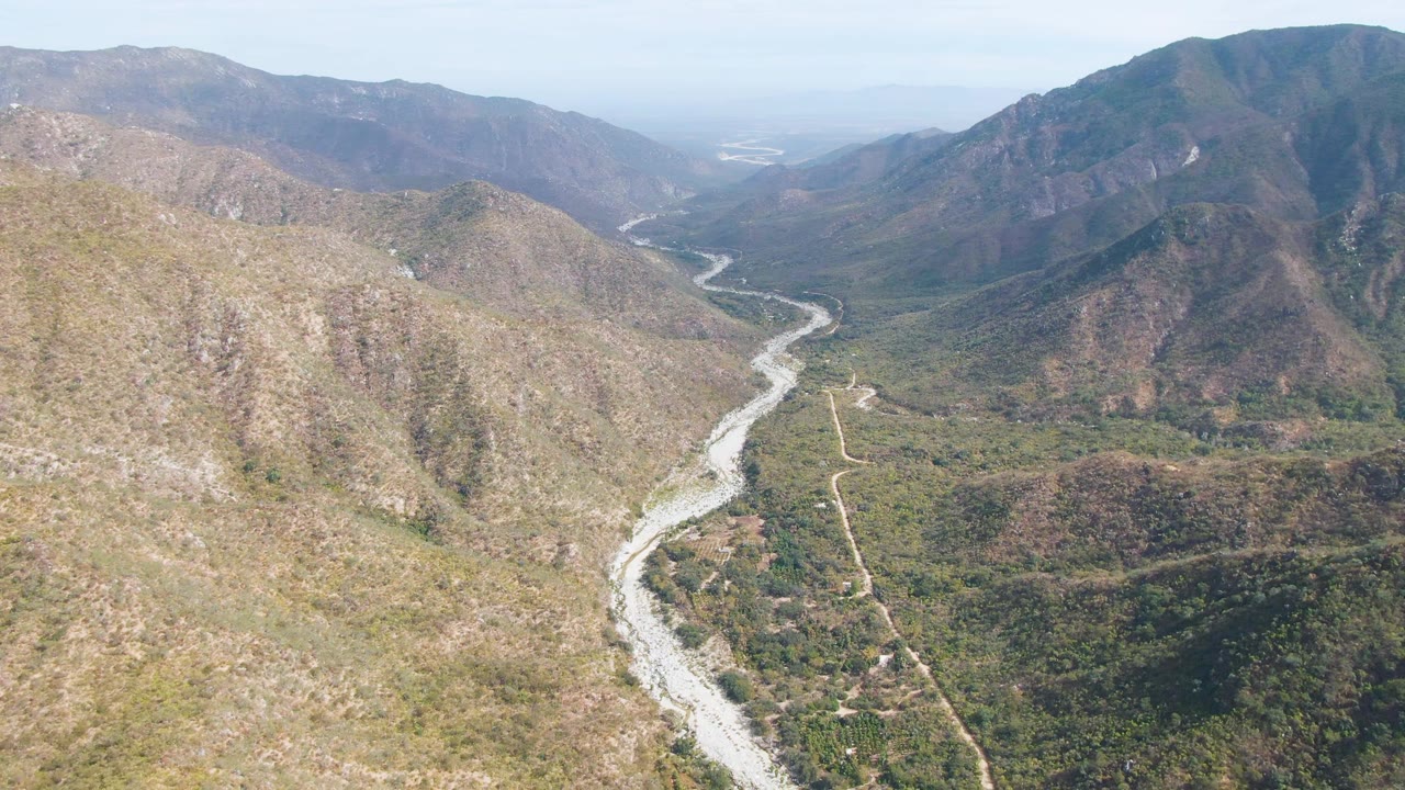 Aerial frame in San Dionisio, Baja California Sur where the riverbed runs from the lower foreground toward distant mountains, with dry slopes, green patches, and no visible people.