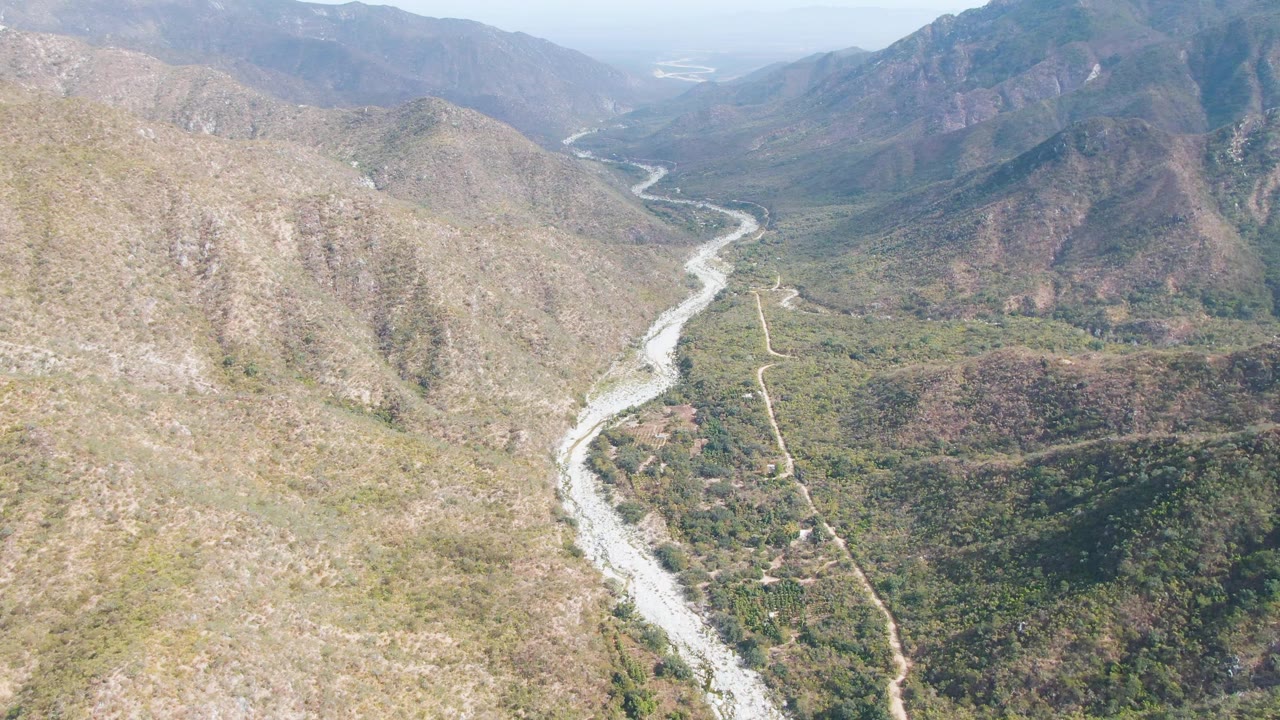 San Dionisio aerial view with a bright riverbed curving away through the valley, dry tan hills on both sides, darker green vegetation patches, a road line, and no visible people.